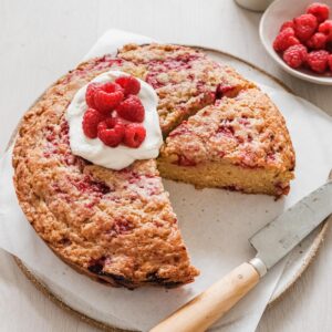 Cake with slices taken decorated with yogurt and raspberries on a plate with a knife and fresh raspberries off to the side.