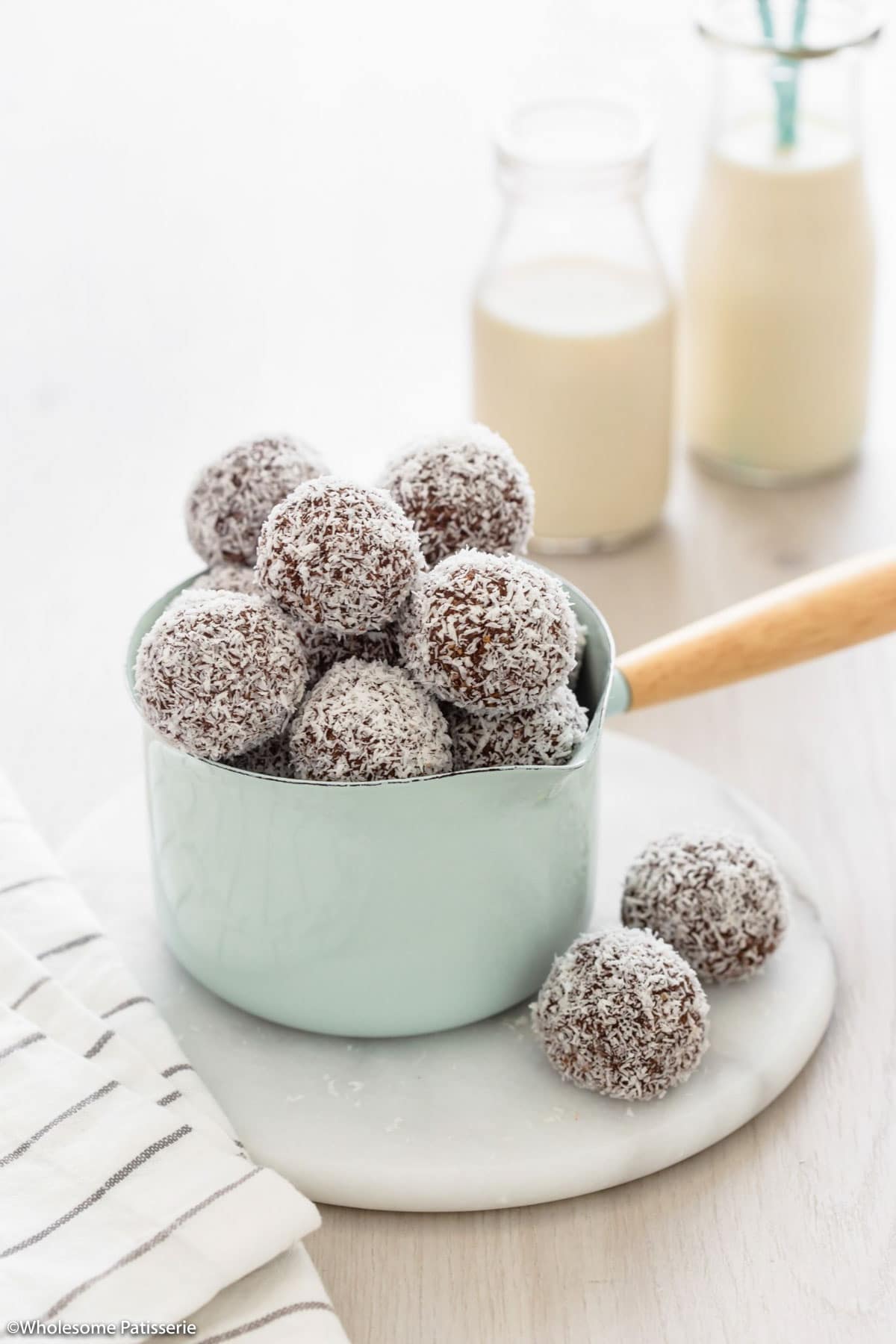 Rum balls without rum coated in coated sitting in green pot on white round platter next to striped tea towel and glasses of milk behind.