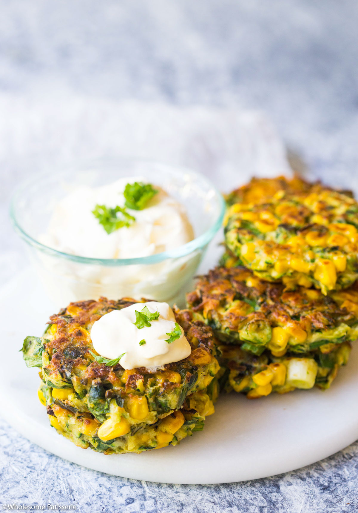 Cooked fritters stacked together on a plate next to a bowl of sour cream.