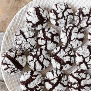Christmas chocolate crinkle cookies on white plate surrounded by more cookies, fairy lights and baubles behind.