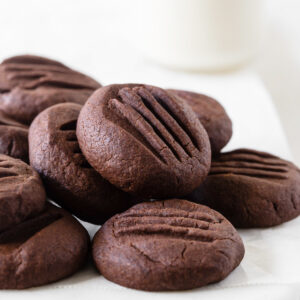 Chocolate meltaway cookies stacked together in a pile on a white platter with a glass of milk behind.