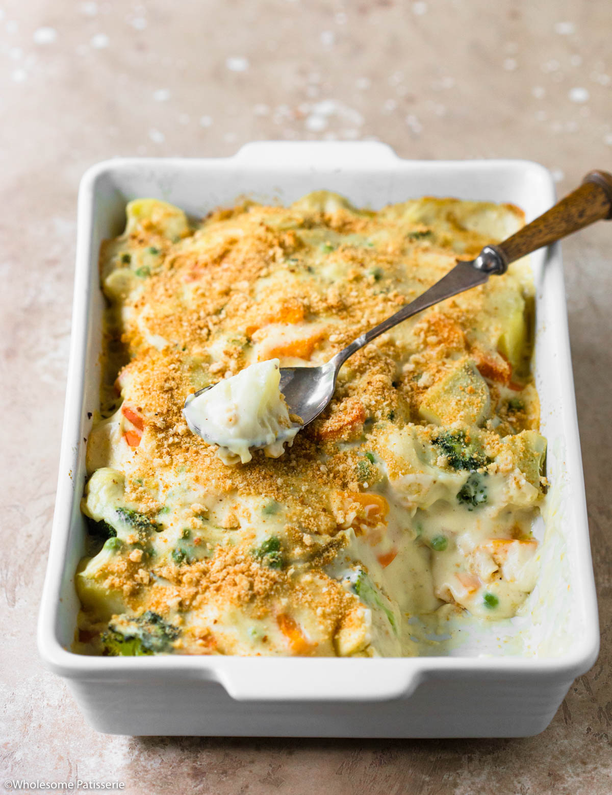 Vegetable bake with white sauce in white baking dish with spoonful taken out sitting on top.