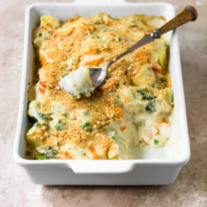 Vegetable bake with white sauce in white baking dish with spoonful taken out sitting on top.