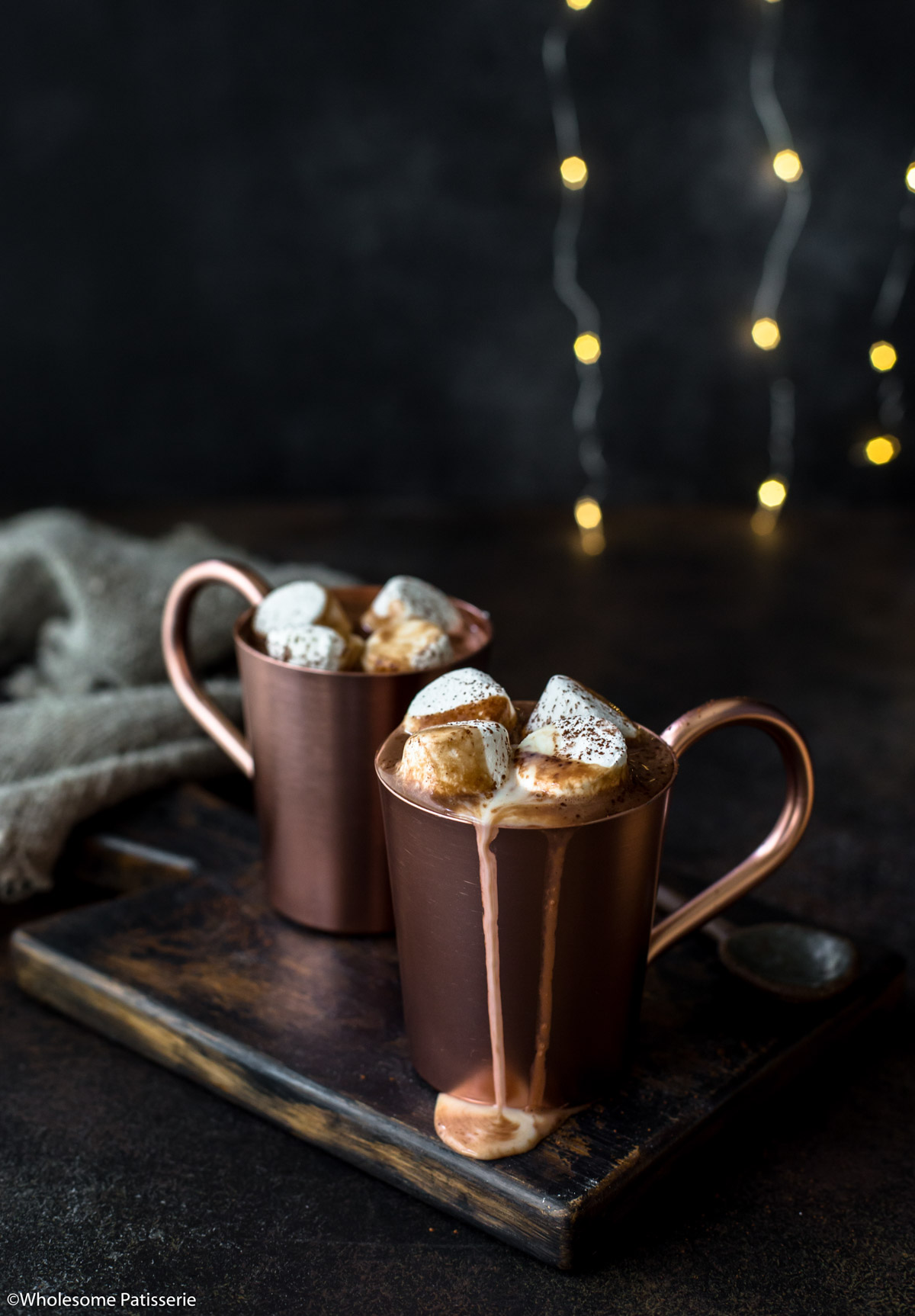 Rum hot chocolate drizzling out of copper mugs with marshmallows and fairy lights behind.