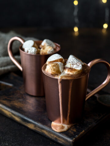 Spiked hot chocolate overflowing out of copper mugs with marshmallows and fairy lights behind.
