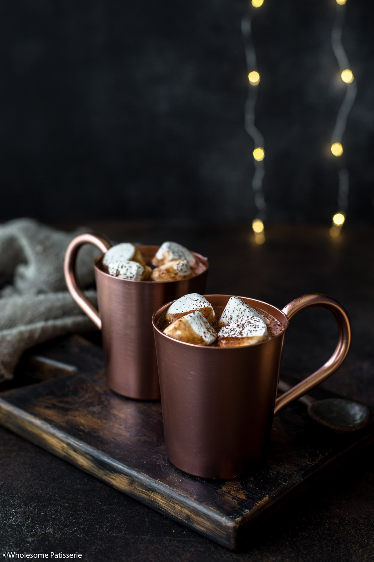 Hot chocolate in two copper mugs topped with marshmallows and cocoa powder with fairy lights behind.