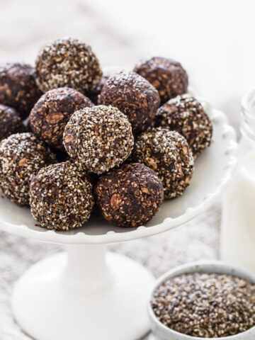 Chia date bliss balls on white stand next to glass of milk and bowl of chia seeds.