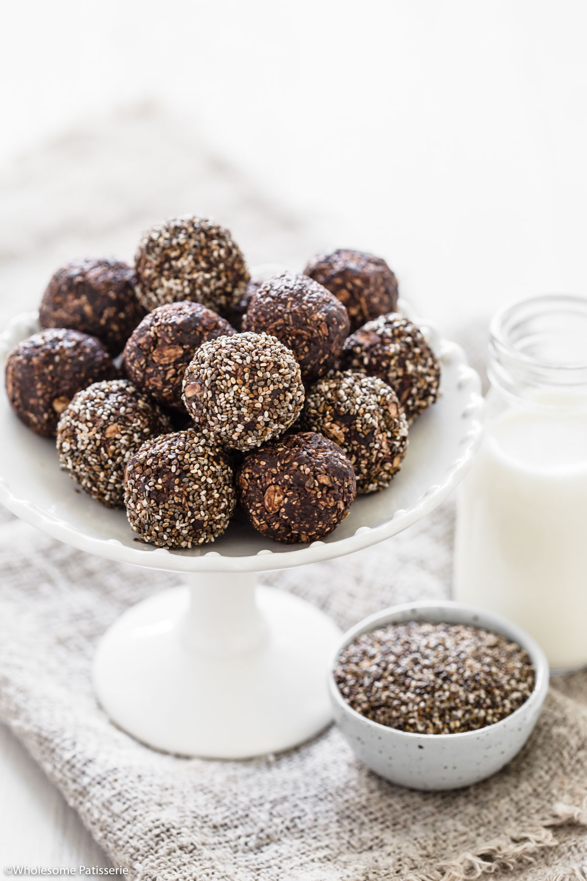 Date bliss balls together on white cake stand next to small bowl of chia seeds and a glass of milk.