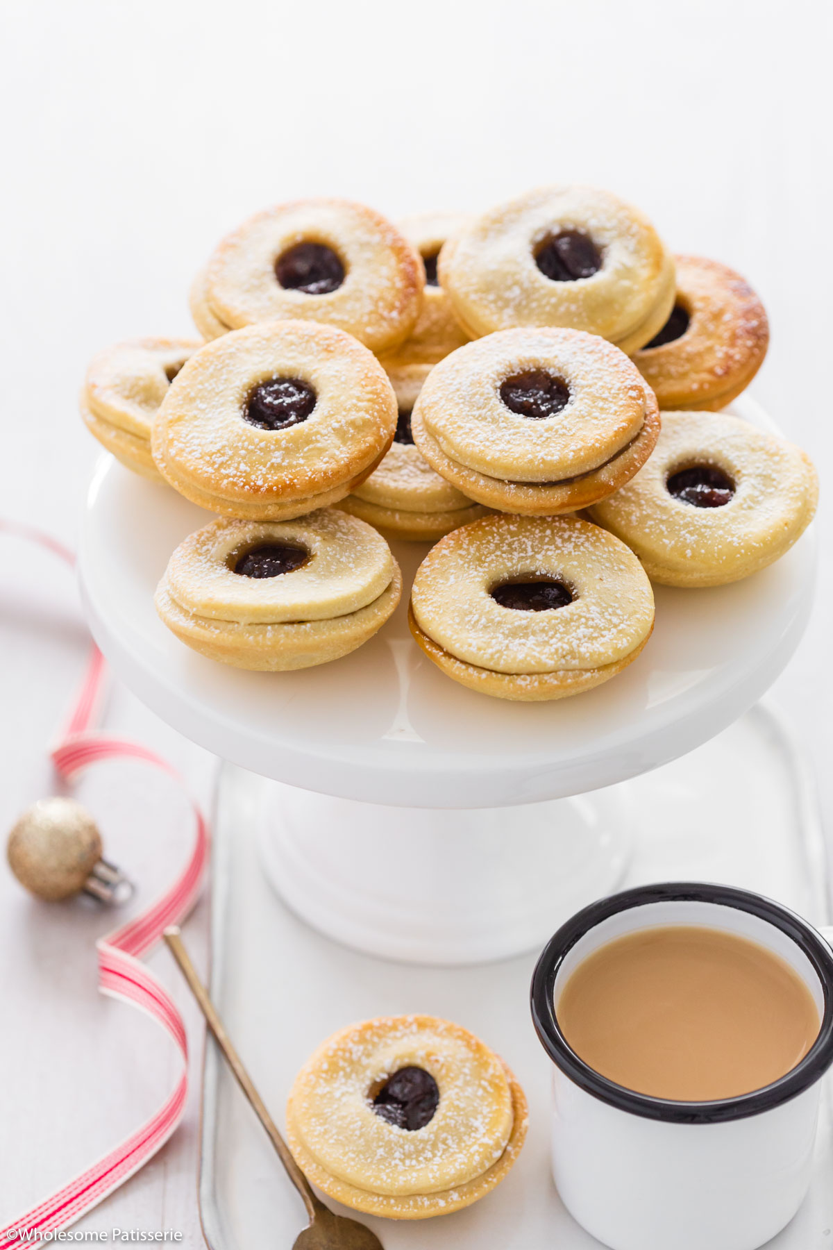 Mince pies on white cake stand next to a cup of tea and red christmas ribbon.