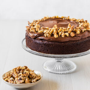 Moist chocolate walnut cake on glass cake stand with a bowl of walnuts in front.