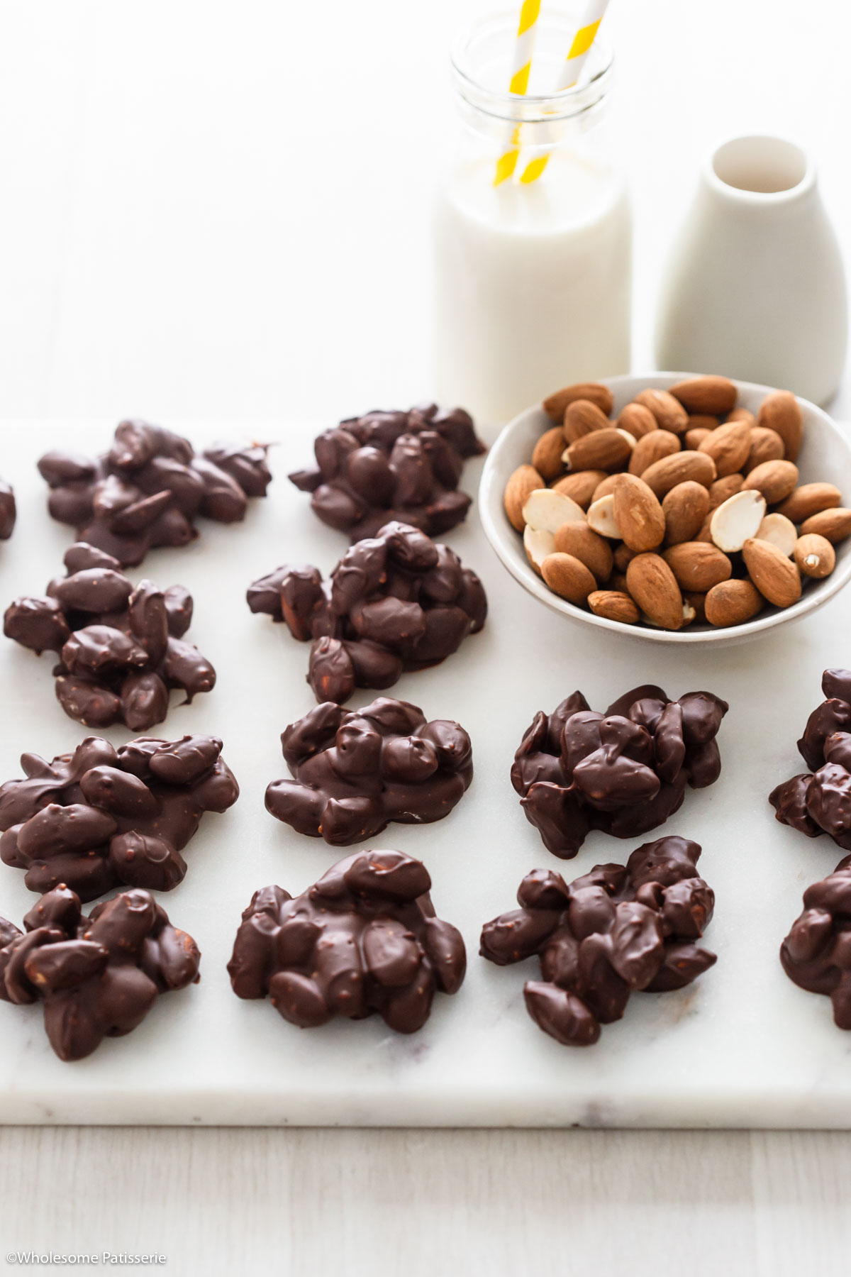 Chocolate covered almonds sitting on white marble platter next to bowl of whole almonds and glass of milk