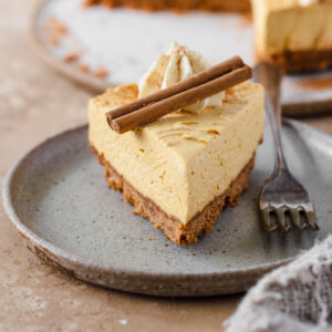 Slice of pumpkin cheesecake on plate next to fork decorated with whipped cream and a cinnamon stick