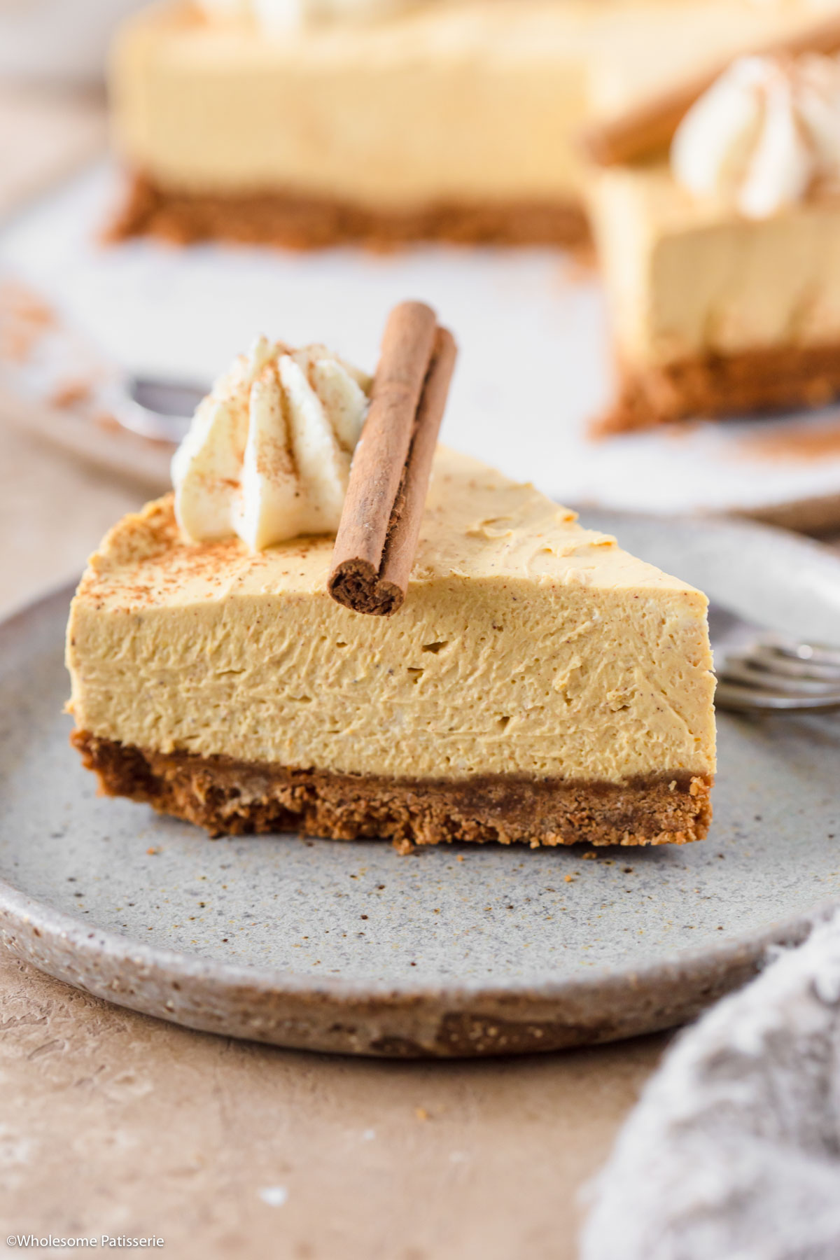 One slice of cheesecake on plate decorated with piped whipped cream, pumpkin pie spice and a cinnamon stick on top