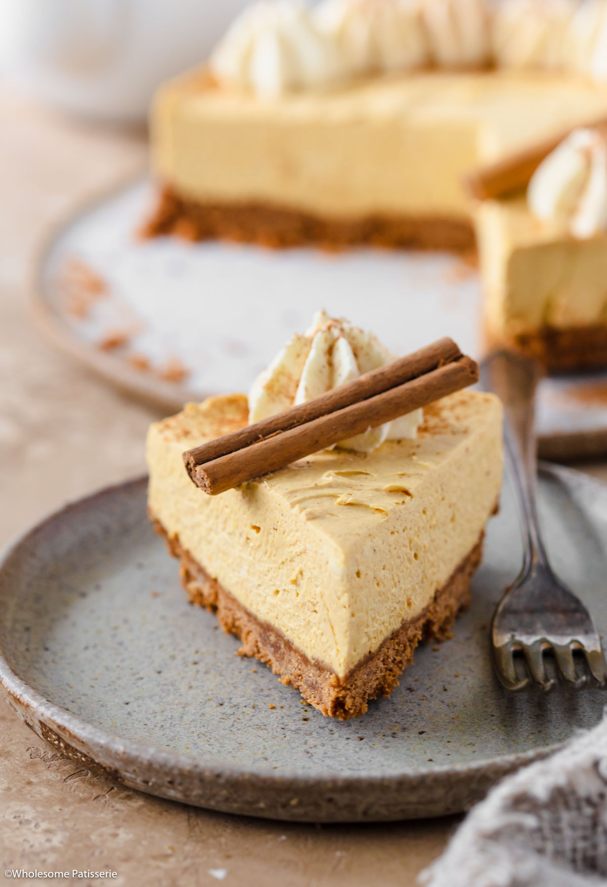 Slice of pumpkin cheesecake on plate next to fork decorated with whipped cream and a cinnamon stick