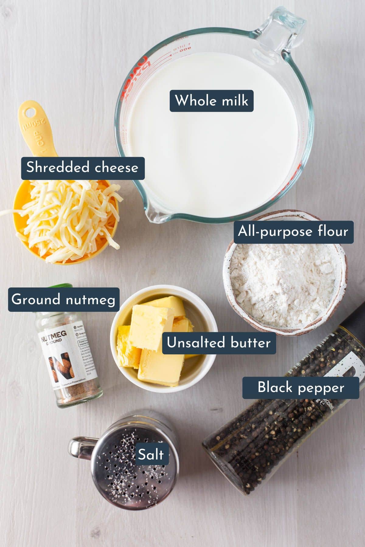 Individual ingredients laid out to make béchamel sauce are whole milk, unsalted butter, all-purpose flour, ground nutmeg, salt and pepper and shredded cheese.