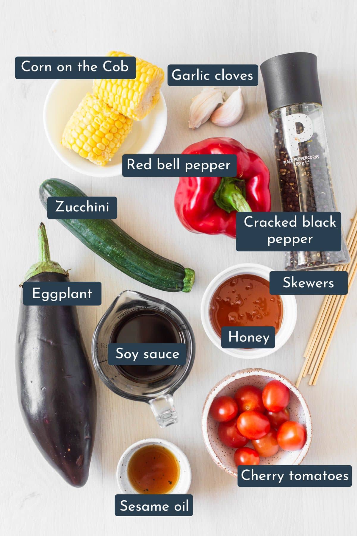 Individual ingredients laid out to make vegetable bbq skewers are garlic, honey, soy sauce, sesame oil, cracked black pepper, zucchini, eggplant, red bell pepper, cherry tomatoes, corn on the cob and wooden or metal skewers.