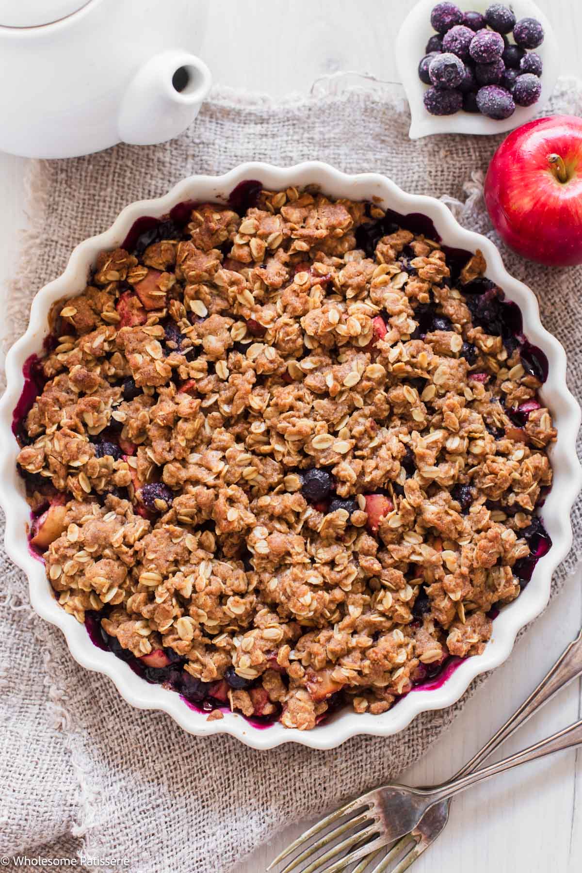 Blueberry apple crumble in white pie dish next to red apple.