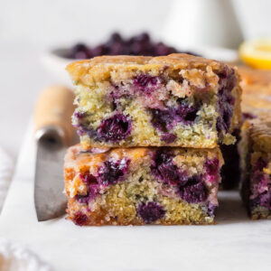 Blueberry cake slices stacked on top of each other on white platter with fresh blueberries and whole lemons behind.