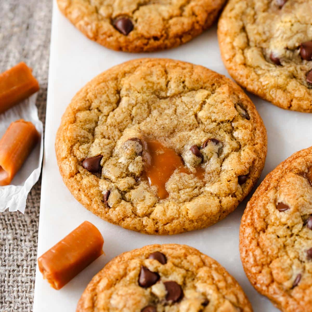 Caramel chocolate chip cookies sitting on white platter with caramel candies unwrapped and sitting around cookies.