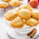 Pies stacked on top of each other sitting on glass stand with apples behind and cinnamon sticks in front