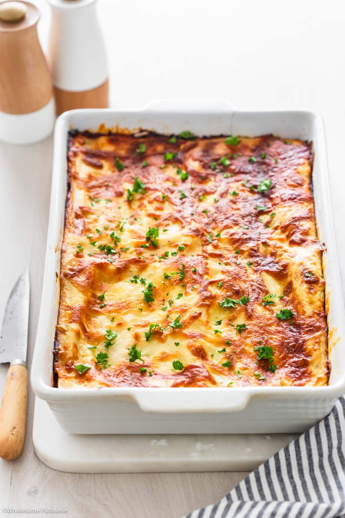 Whole vegetable tomato lasagna in baking dish displayed on white board next to wooden knife.