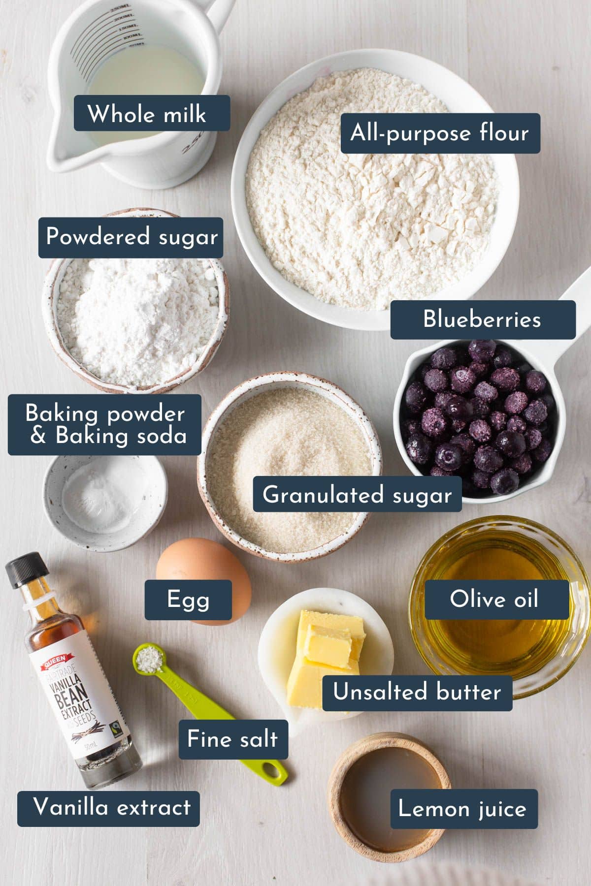 Individual ingredients laid out to make moist blueberry cake with lemon glaze are all-purpose flour, whole milk, baking powder, baking soda, salt egg, granulated sugar, olive oil, vanilla extract, blueberries, unsalted butter, lemon juice and powdered sugar.