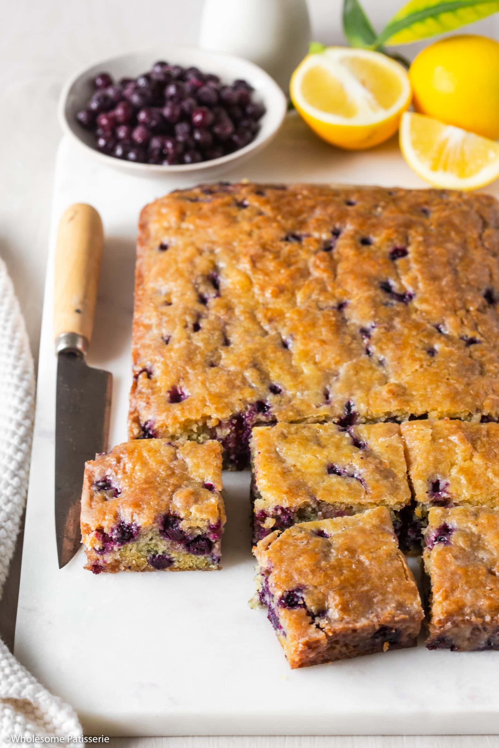 Blueberry cake sliced into squares sitting on white platter next to brown handle knife with whole fresh lemons and blueberries behind.