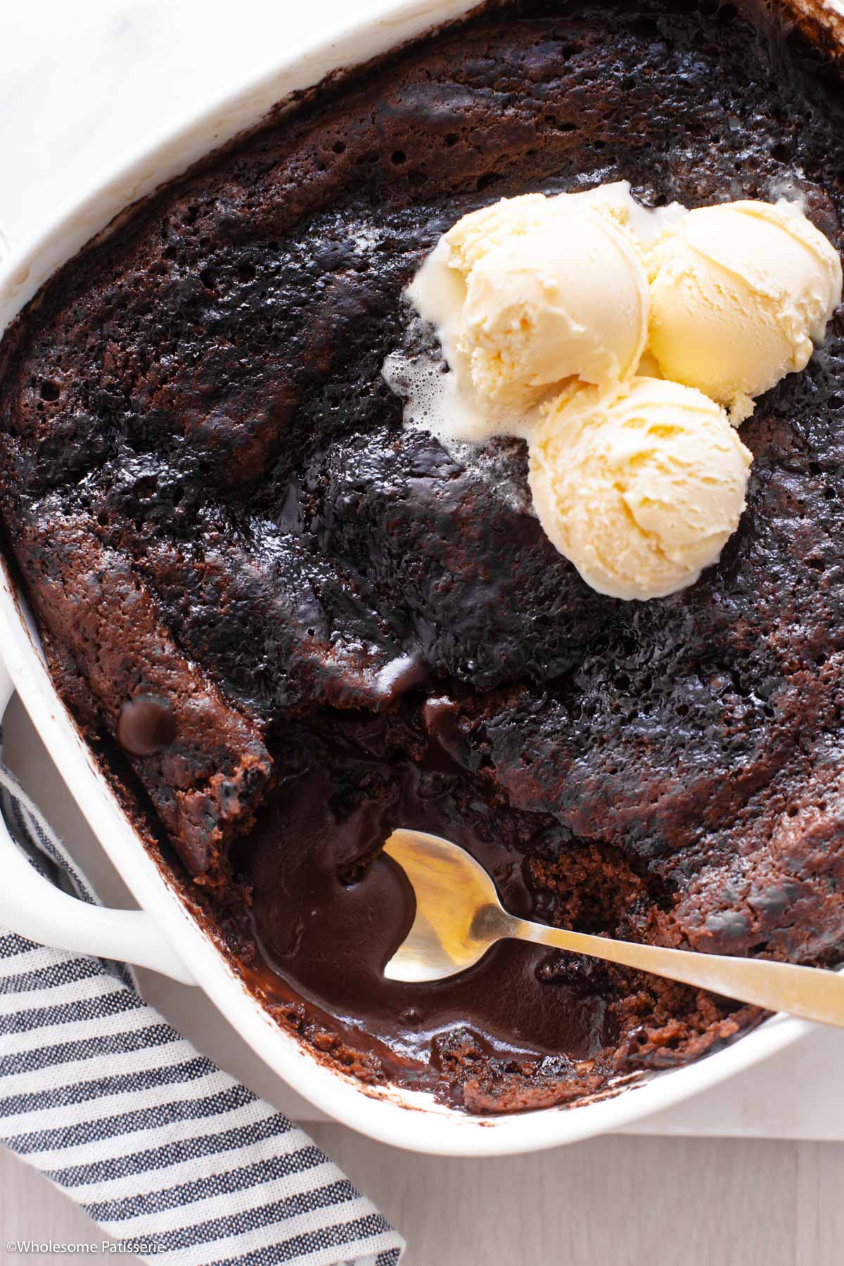 Overhead view of chocolate self saucing pudding in baking dish with gold spoon and scoops of vanilla ice cream