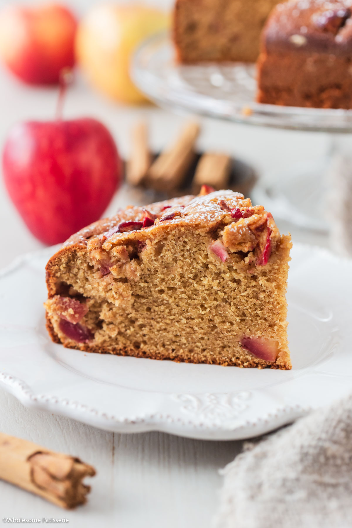 One slice of cinnamon apple cake on white plate with red apple and cinnamon sticks behind.
