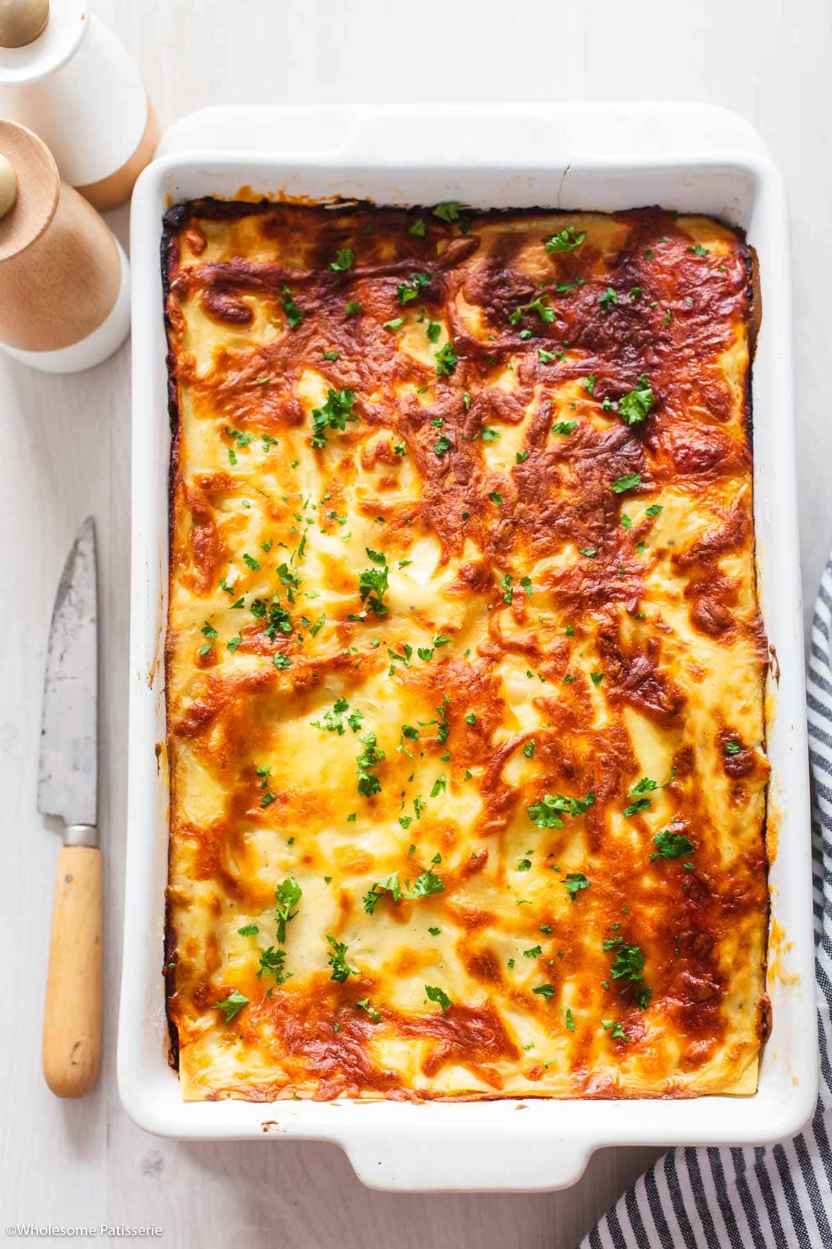 Overhead view of vegetable lasagna with tomato in white baking dish garnished with chopped parsley next to wooden knife.