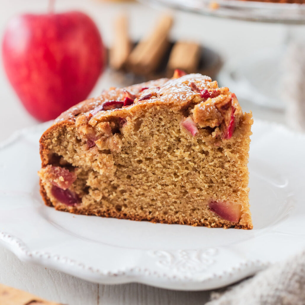Slice of cinnamon apple cake on white plate with red apple behind.