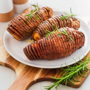 Hasselback sweet potatoes on serving plate sitting on a large wooden board and garnished with fresh rosemary sprigs.