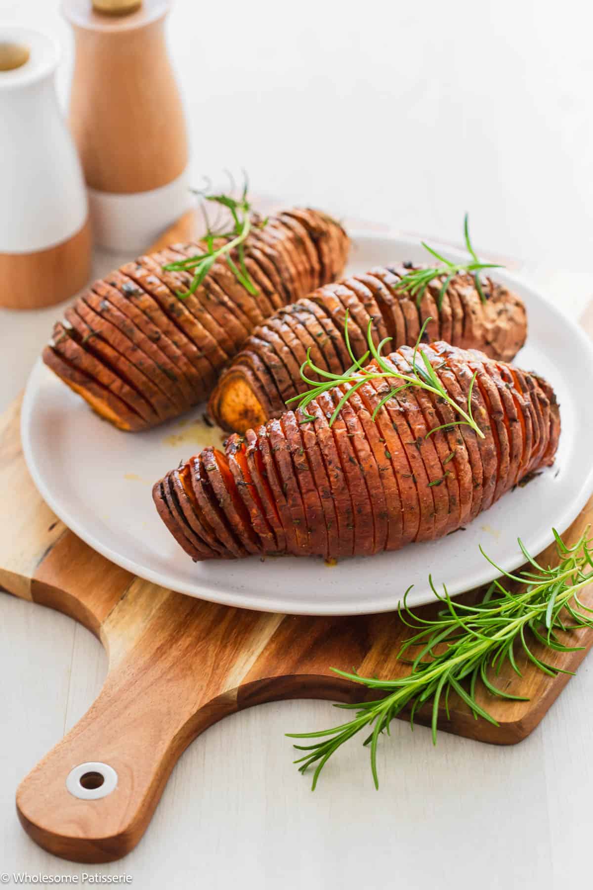 Hasselback sweet potatoes on serving plate sitting on a large wooden board and garnished with fresh rosemary sprigs.