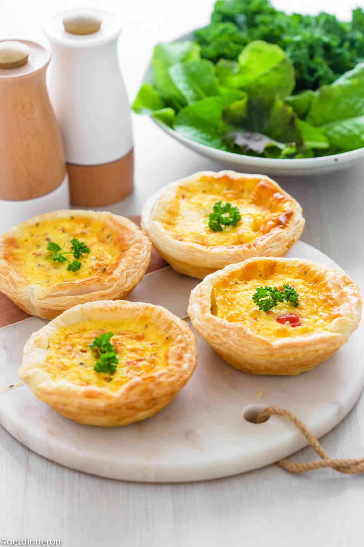 Individual quiches garnished with fresh parsley and sitting on marble platter with bowl of leafy green salad behind.