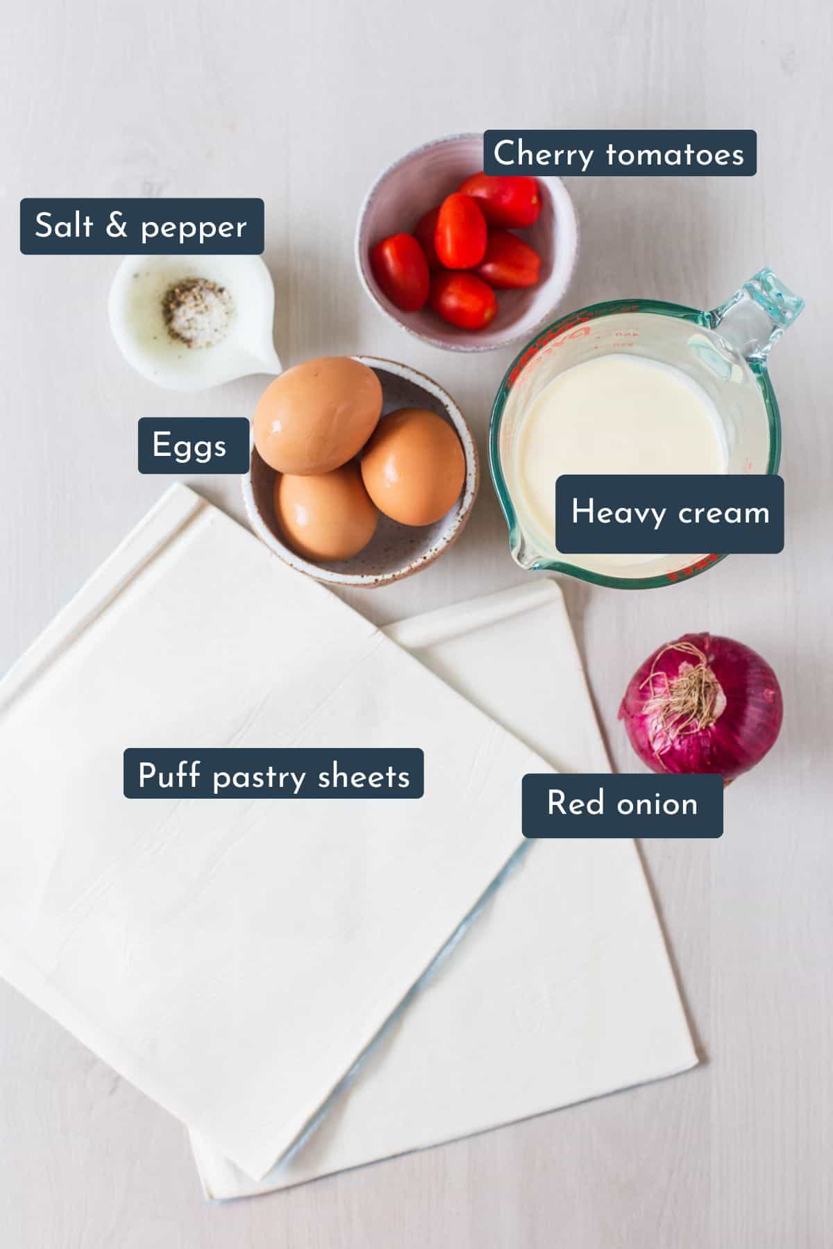 Individual ingredients laid out to make puff pastry quiches are puff pastry sheets, red onion, eggs, heavy cream, cherry tomatoes, salt and pepper.