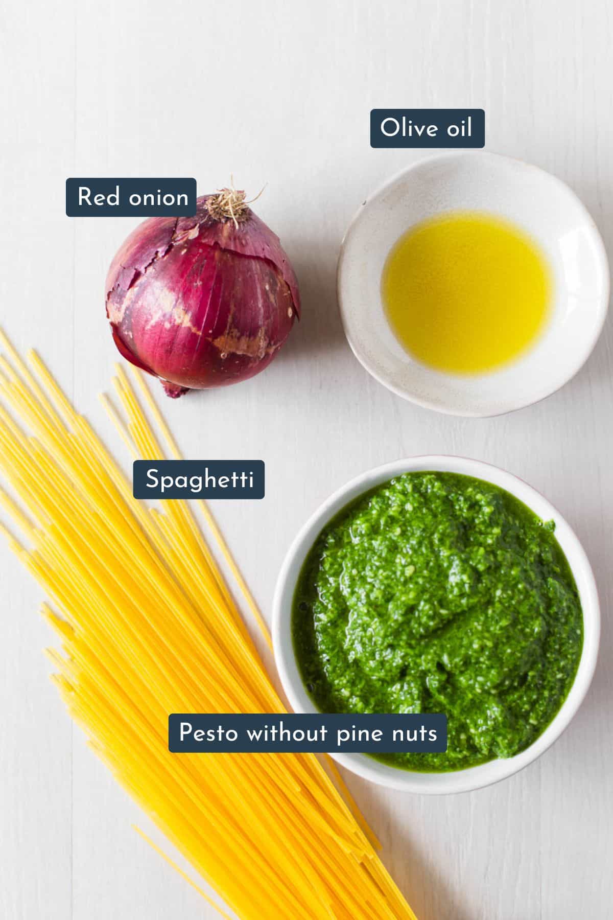 Individual ingredients laid out to make pasta al pesto are spaghetti, pesto sauce, red onion and oil.