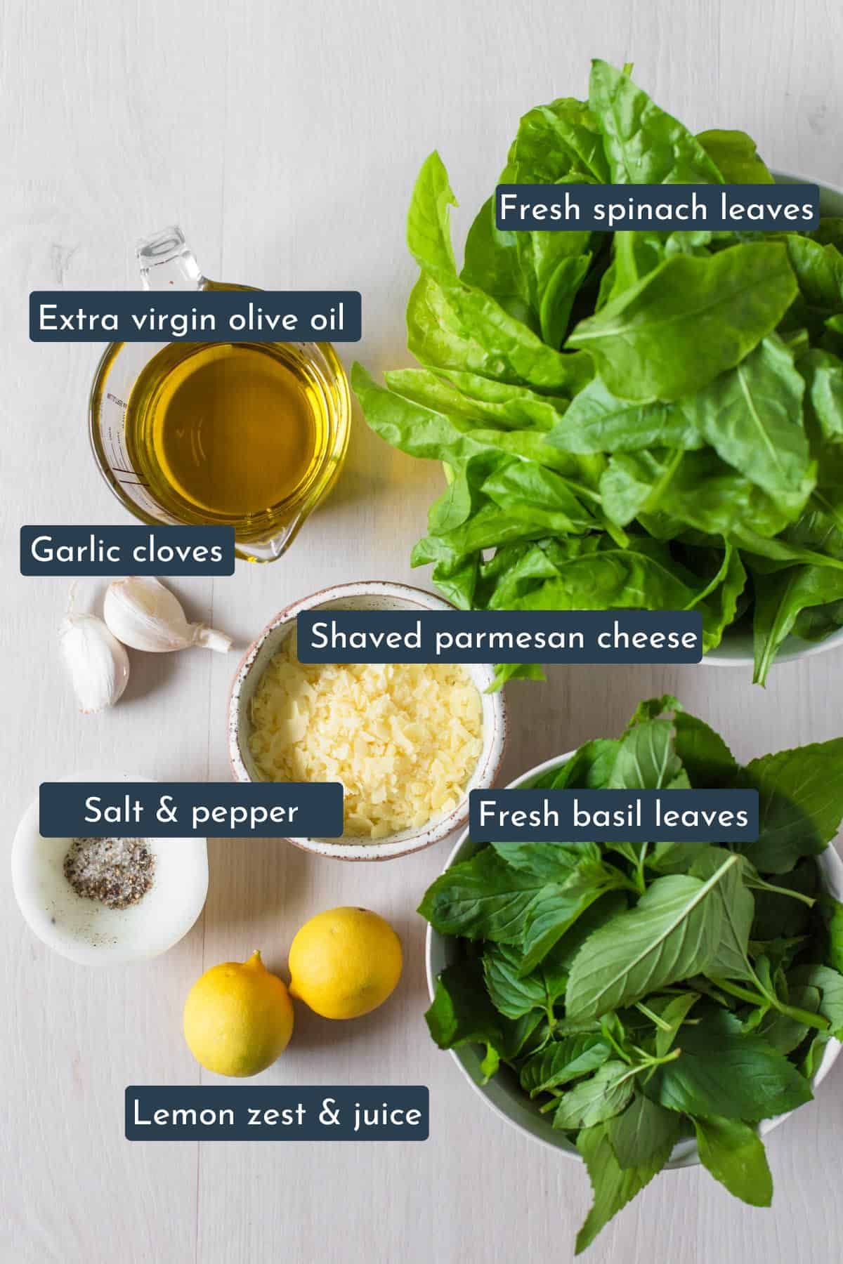 Individual ingredients laid out to make pesto without pine nuts are spinach, basil, garlic cloves, olive oil, lemon, salt, pepper and parmesan cheese.