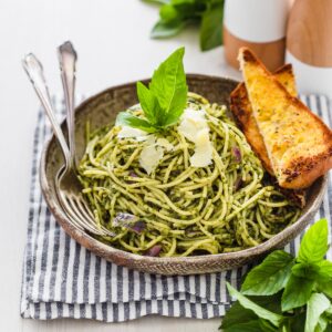 Pasta al pesto served in bowl garnished with shaved parmesan and fresh basil with a side of crunchy bread.