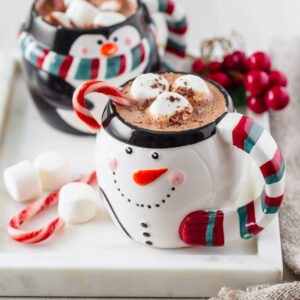 Peppermint hot chocolate in two christmas themed mugs with candy canes and marshmallows next to them.
