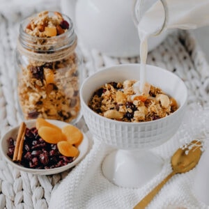 Milk poured into bowl with granola and dried fruit in bowls behind.