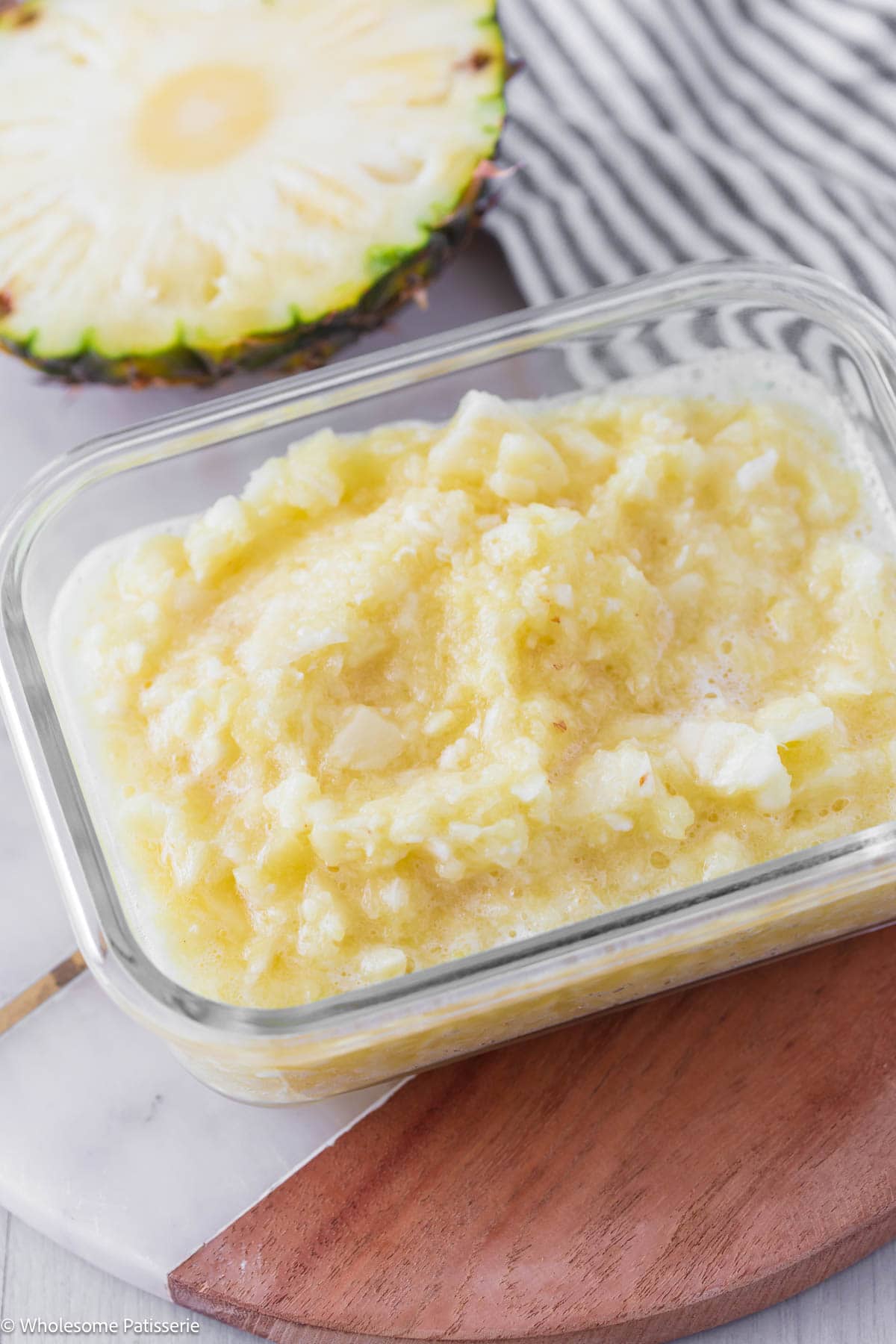 Crushed pineapple in glass container with fresh pineapple behind.