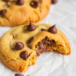 Chocolate chip cookie with bite taken out and sitting on white parchment paper.
