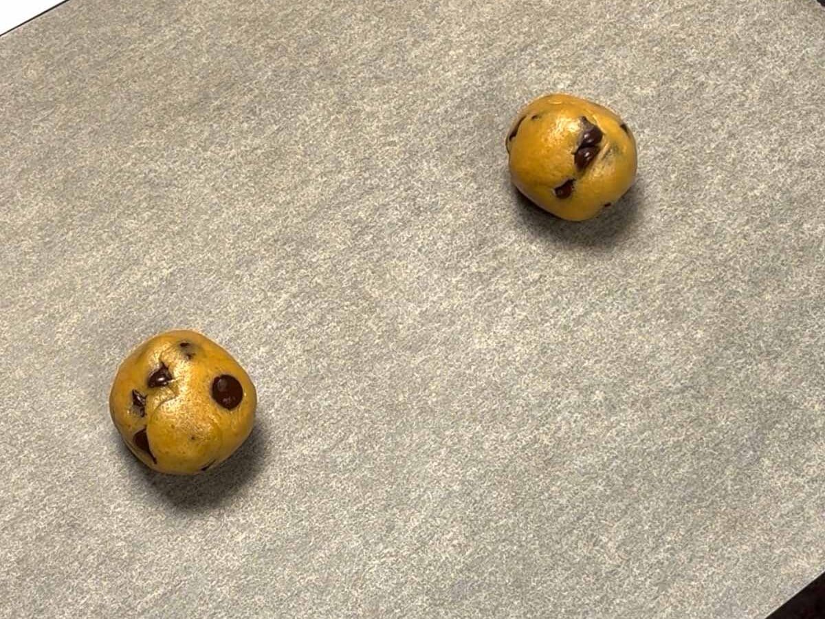 Two chocolate chip cookie dough balls on baking sheet before baking.