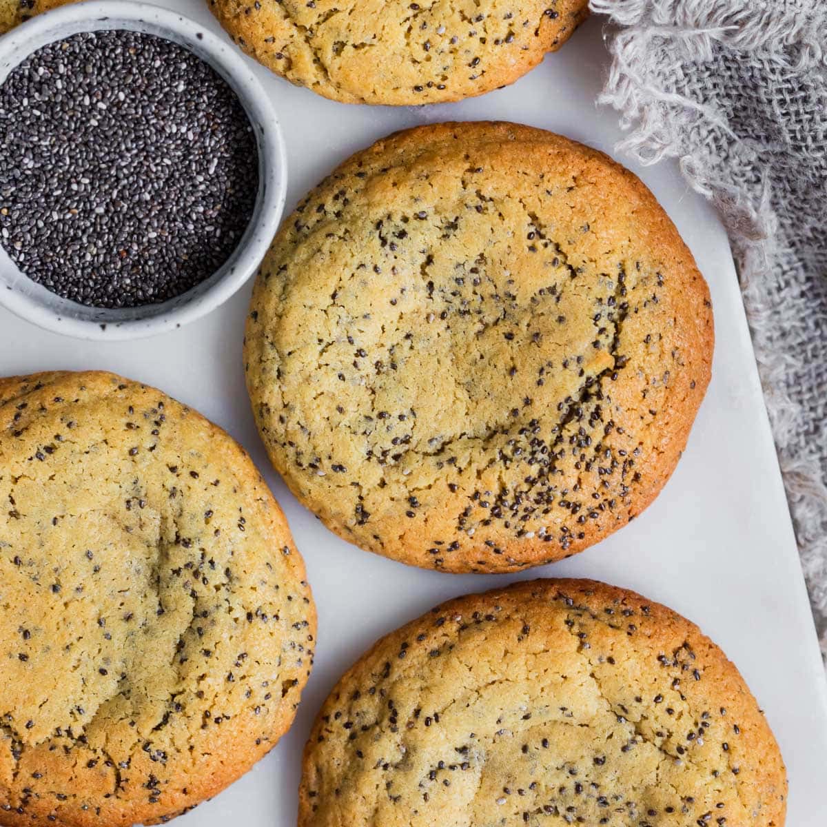 Big soft chia seed cookies on marble board with small bowl of chia seeds.