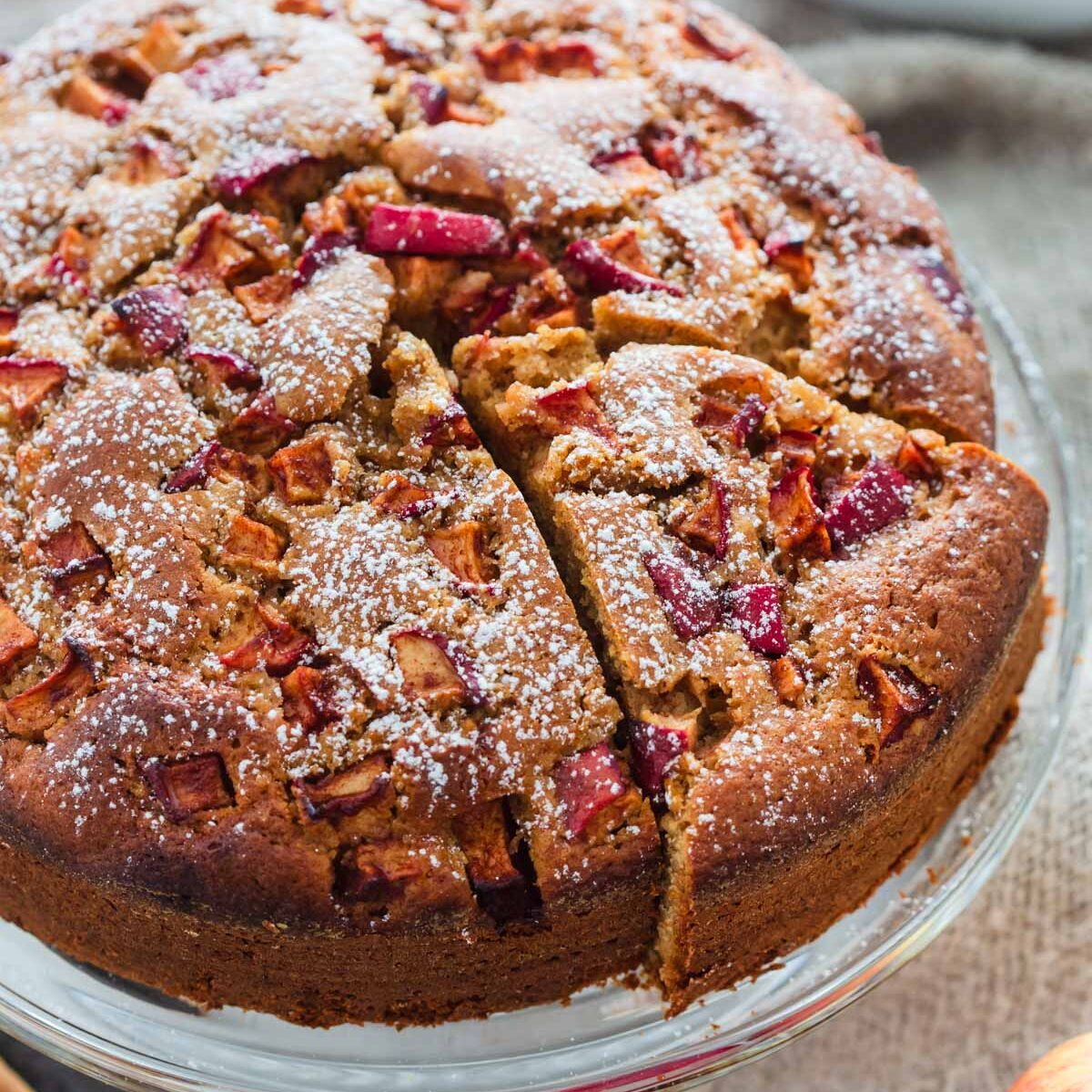 Cinnamon apple cake dusted with powdered sugar on glass cake stand with slice cut out of it.
