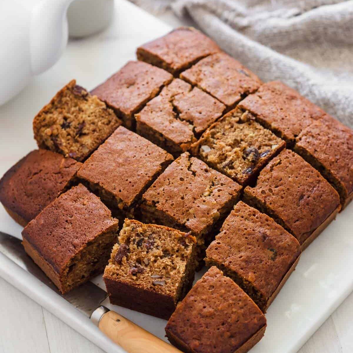 Slab of baked raisin cake cut into squares and sitting on white marble board next to knife.