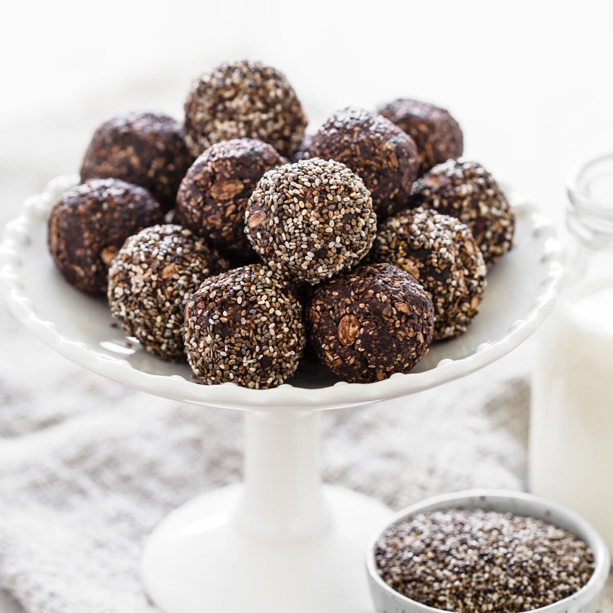 Chia date bliss balls on white cake stand next to glass of milk and bowl of chia seeds.